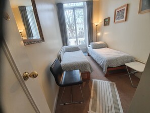 Ein kleiner, gut beleuchteter Raum mit zwei Betten, einem Sessel, einem Heizkörper und einem Fenster mit Vorhängen.