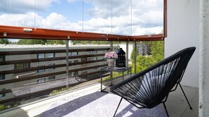 Un balcón con una silla, una mesita con un florero y una decoración con forma de casa, y vistas a edificios y árboles.