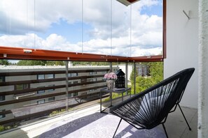 Uma varanda com cadeira, uma mesa pequena com um vaso e uma decoração em formato de casa, e vista para edifícios e árvores.