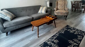 Una sala de estar con un sofá gris, una mesa de centro de madera, un sillón estampado, un ventilador de techo y un tapete oscuro con un diseño de árbol.