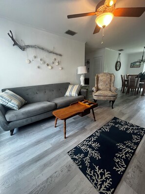 Ein Wohnzimmer mit grauem Sofa, einem Holztisch, einem gemusterten Sessel, einem Deckenventilator und einem dunklen Teppich mit Baumdekor.
