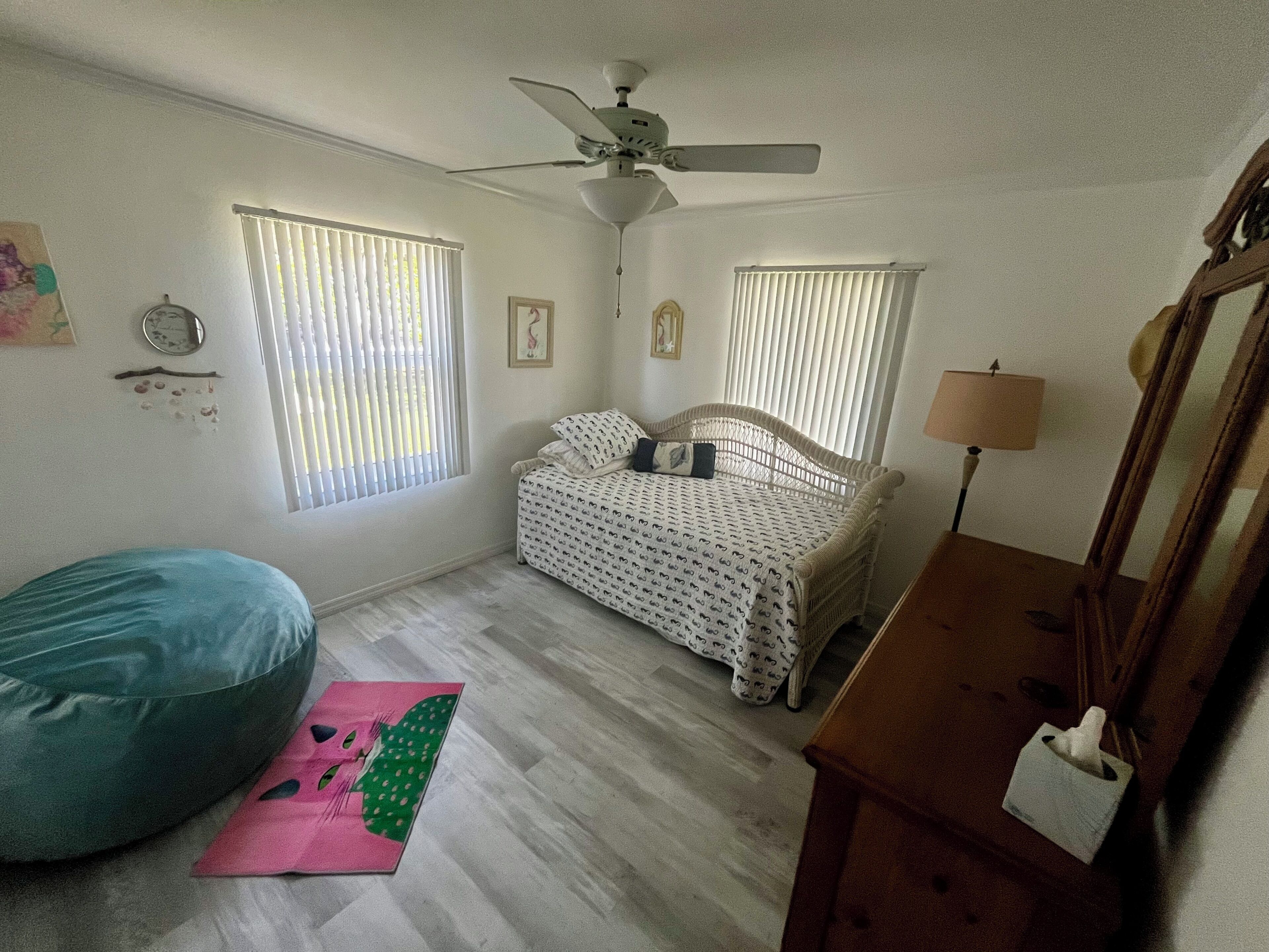 A bedroom with a bed, a ceiling fan, two windows with blinds, a bedside lamp, a bedside table with a tissue box, a bean bag chair, and a rug.