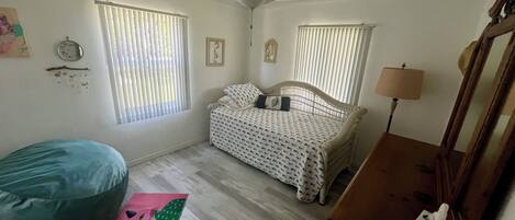 A bedroom with a bed, a ceiling fan, two windows with blinds, a bedside lamp, a bedside table with a tissue box, a bean bag chair, and a rug.