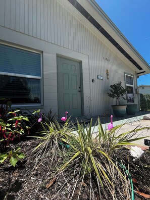 A building with a grey exterior, a green door, and a window with blinds. There are two large planters with plants and a pink flamingo decoration. - Charming 3-bedroom house in Fort Myers hot tub, fully fenced, close to Sanibel (Fort Myers)