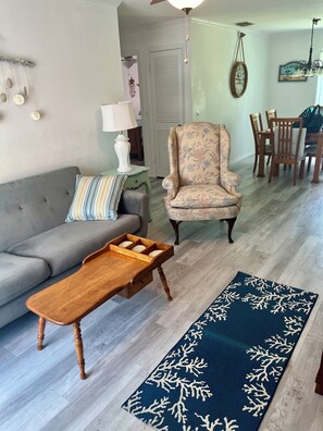 Un salon comprenant un canapé, un fauteuil à motifs, une table basse en bois avec des tiroirs, un tapis bleu orné d’un motif d’arbre blanc, et un coin repas avec des chaises et une table.