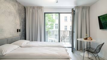 Ein modernes Hotelzimmer mit einem Bett, einem kleinen Tisch mit Flasche und Gläsern, einem Sessel und einem Balkon mit Blick auf Gebäude und Bäume.