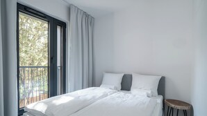 A bedroom with a bed, a small stool, a window with curtains, and a view of trees. - RentPlanet - Jodłowa Residence (Wrocław)