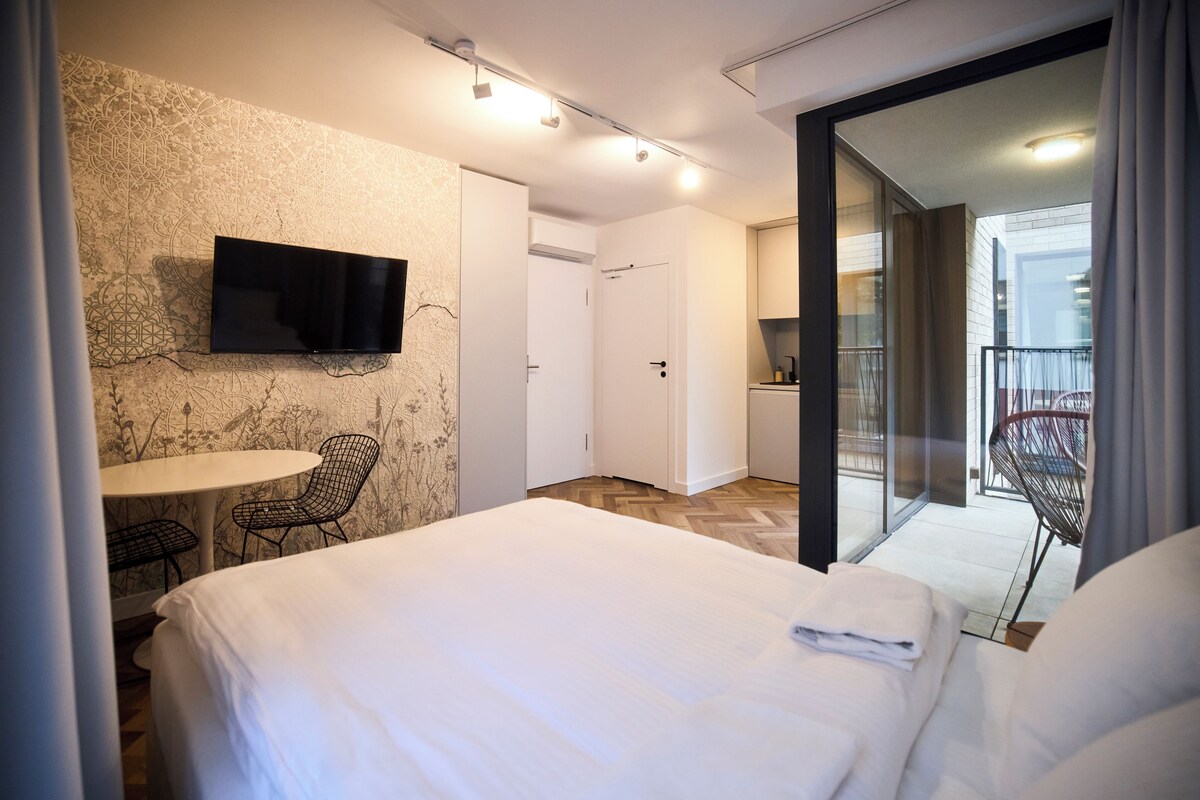 Habitación de hotel con cama, televisor montado en la pared, una silla y balcón con un sillón.