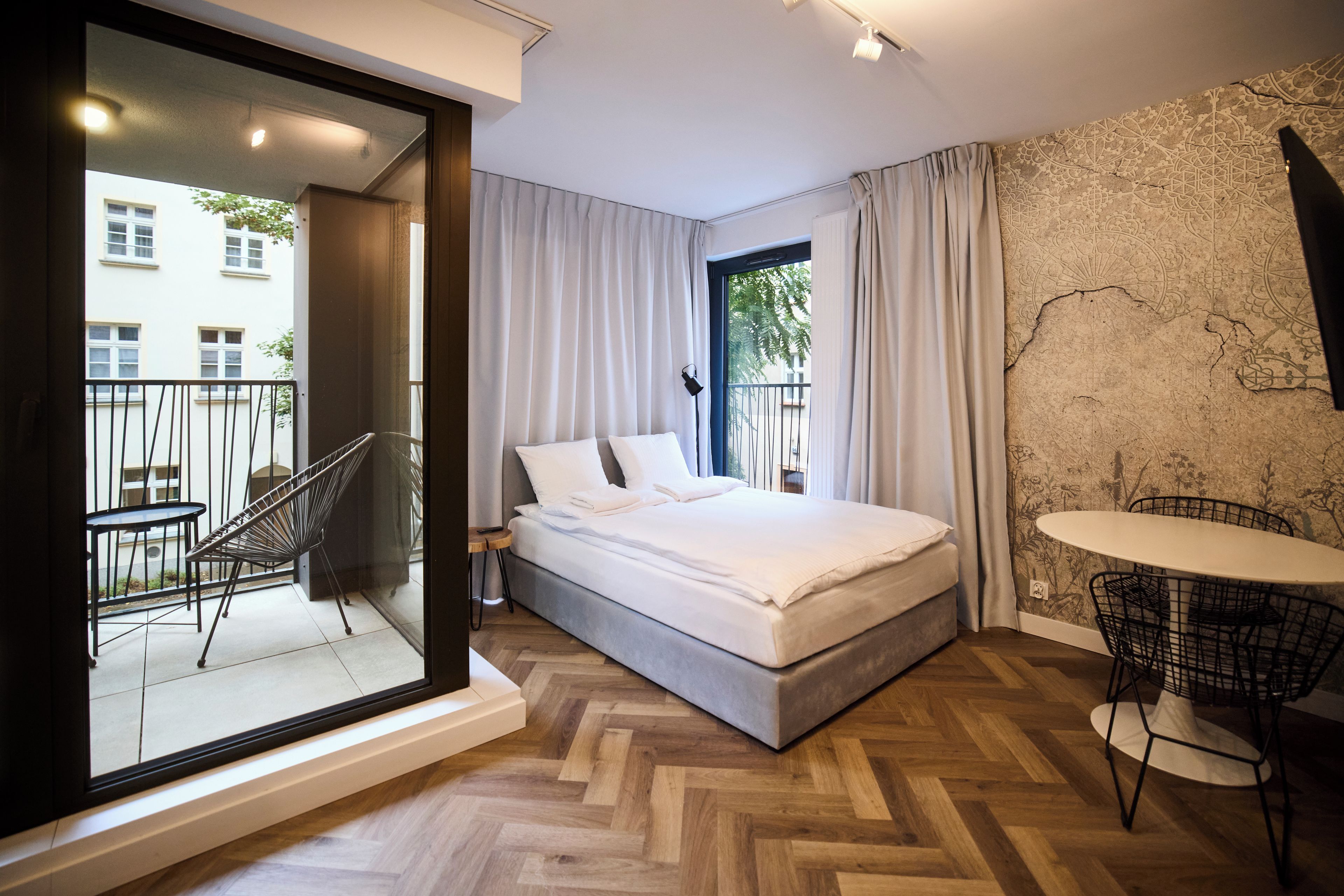 Ein modernes Schlafzimmer mit einem Bett, einem kleinen Tisch und einem Sessel. Ein Balkon mit Geländer bietet Blick auf die umliegenden Gebäude.