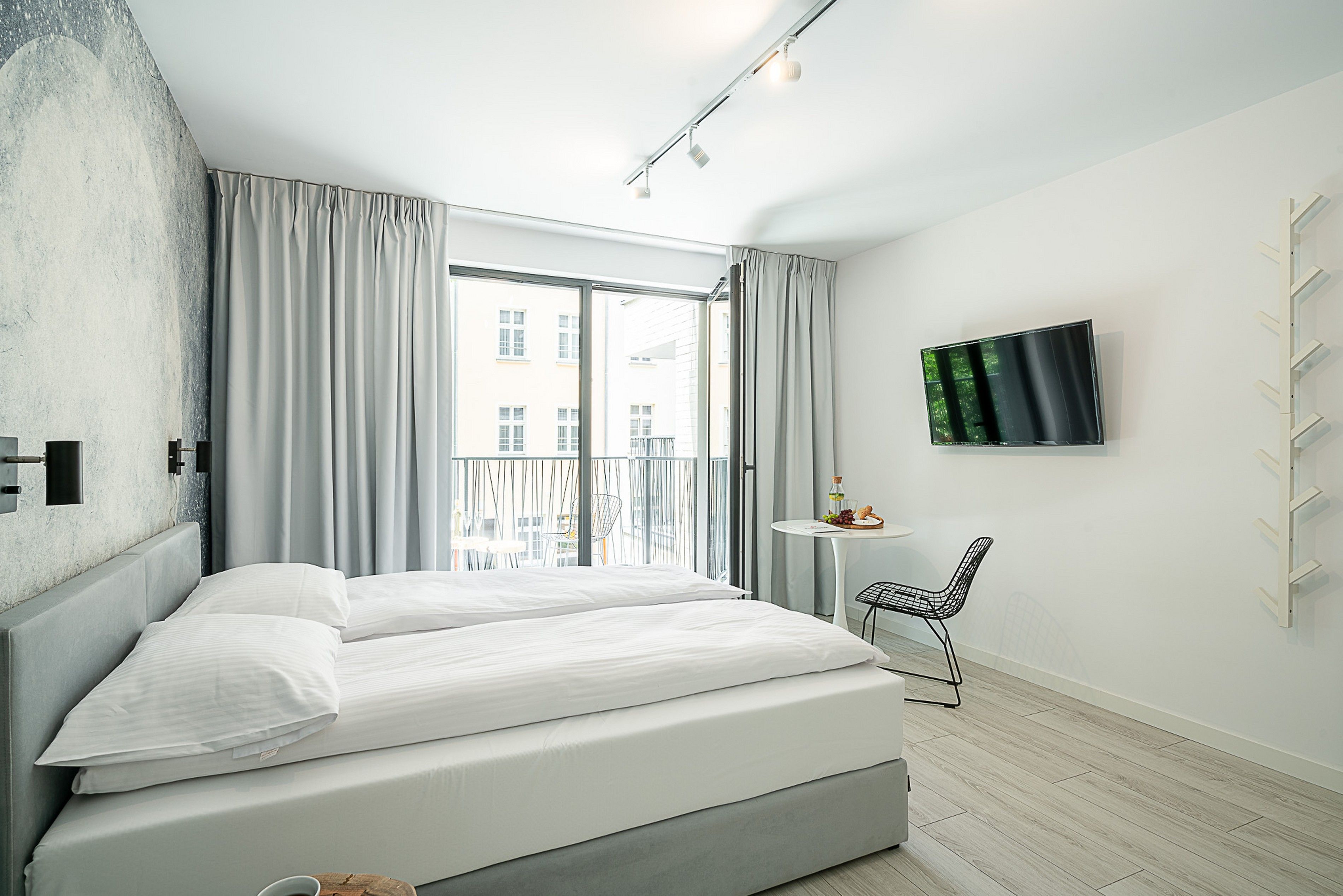 A modern bedroom with a bed, a small table with a chair, a TV, and a balcony with curtains.