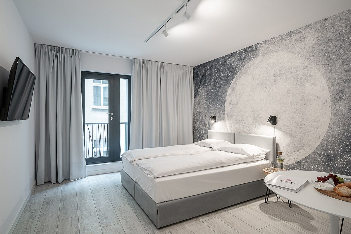 Un dormitorio moderno con cama, televisor montado en la pared, mesita de noche con una botella y vasos, y vistas al exterior.