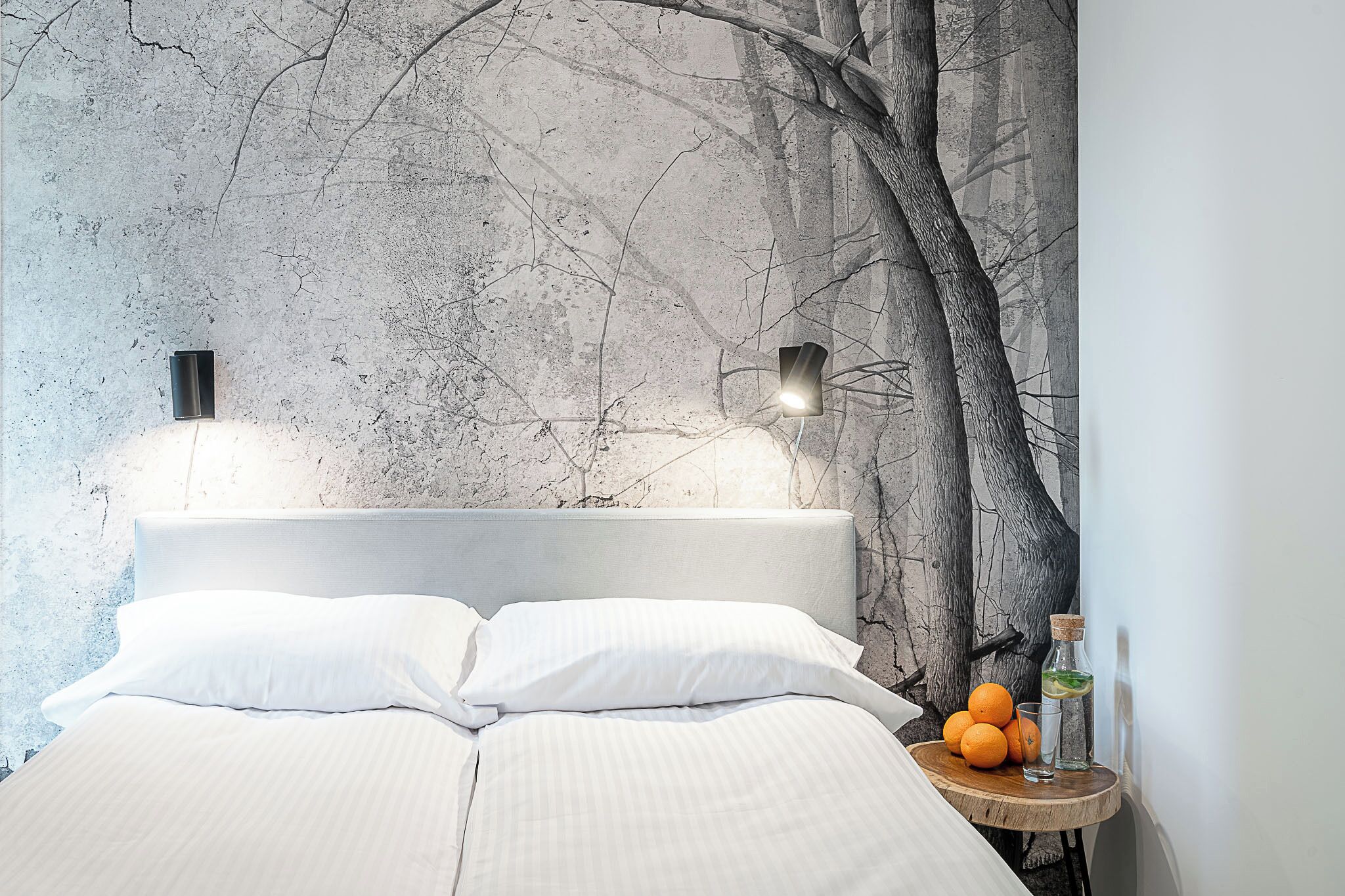 Ein Bett mit weißer Bettwäsche, ein Nachttisch mit einem Glas Wasser und einer Flasche sowie eine Wand mit einer Baum-Mural.