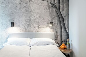 A bed with white linens, a nightstand with a glass of water and a bottle, and a wall with a tree mural. - RentPlanet - Jodłowa Residence (Wrocław)