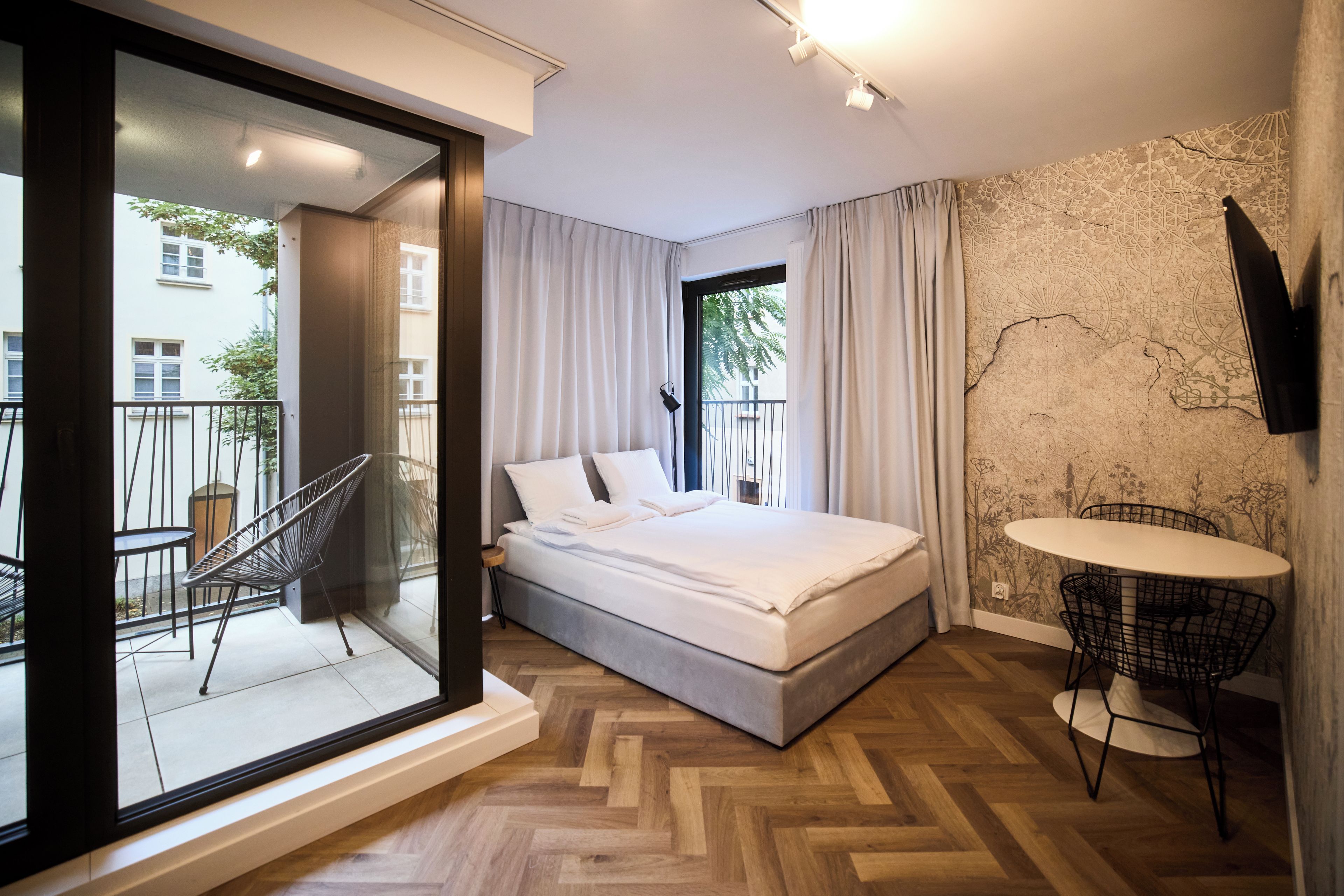 Un dormitorio moderno con cama, un balcón con una silla, una mesita y un televisor montado en la pared.