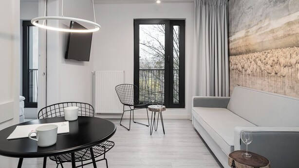 A modern living room with a dining area, a sofa, a TV mounted on the wall, and a large window with curtains. - RentPlanet - Jodłowa Residence (Wrocław)