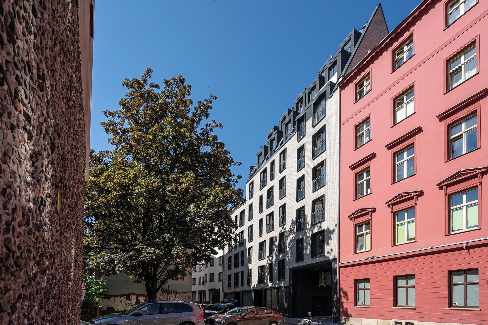 Straßenansicht mit mehrstöckigen Gebäuden, einem Baum und parkenden Autos.