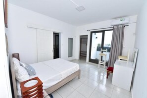 Ein Schlafzimmer mit einem Bett, einem Schreibtisch und einem Stuhl. Eine Tür führt in einen anderen Raum, ein Fenster mit Vorhängen ist ebenfalls vorhanden.