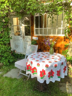 A rustic outdoor seating area with a table covered in a floral tablecloth, a white chair, and a sign reading 'WEJSIE' on a wooden wall.