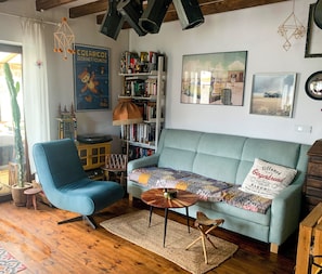 A cozy living room with a blue armchair, a light green sofa, a wooden coffee table, and a bookshelf filled with books and decorative items.