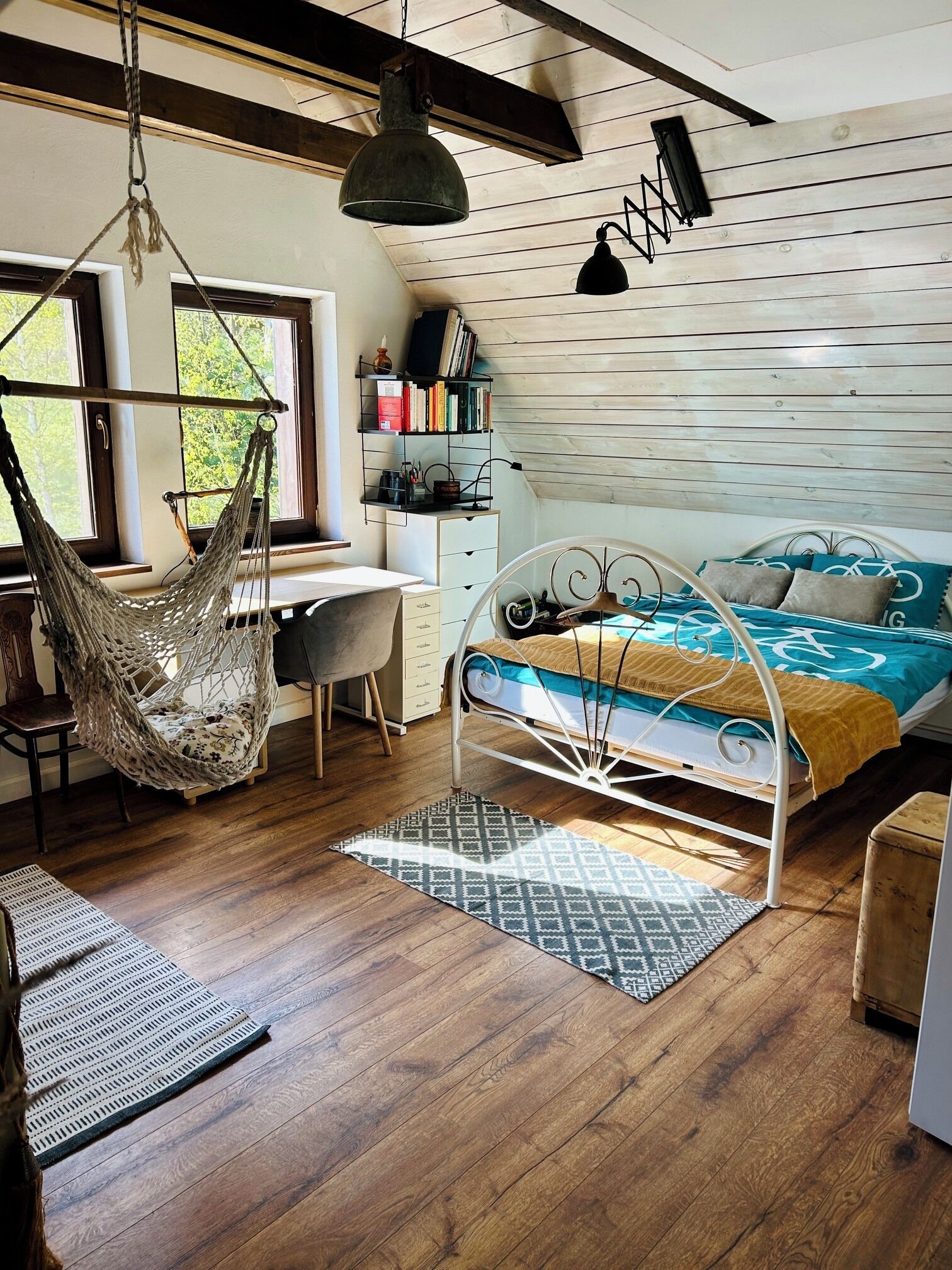 Une chambre confortable avec un hamac, un lit recouvert d’une couvre-lit sur le thème du vélo, un bureau avec des livres et un luminaire suspendu.