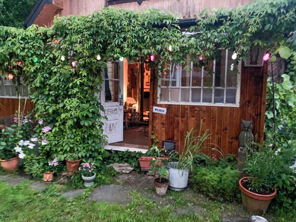 A wooden cottage with a garden entrance, a sign reading 'WEJSIE,' and various potted plants.