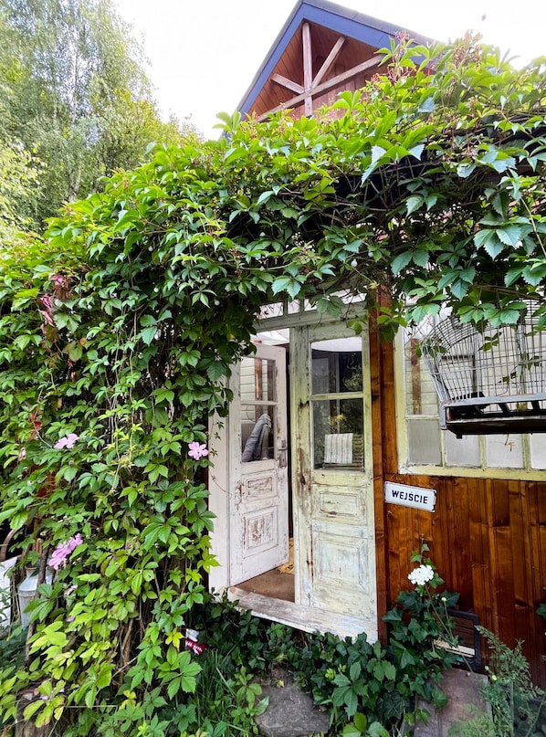 Un bâtiment rustique en bois, avec une porte portant l’inscription « WEJSIE » et une entrée recouverte de vigne.