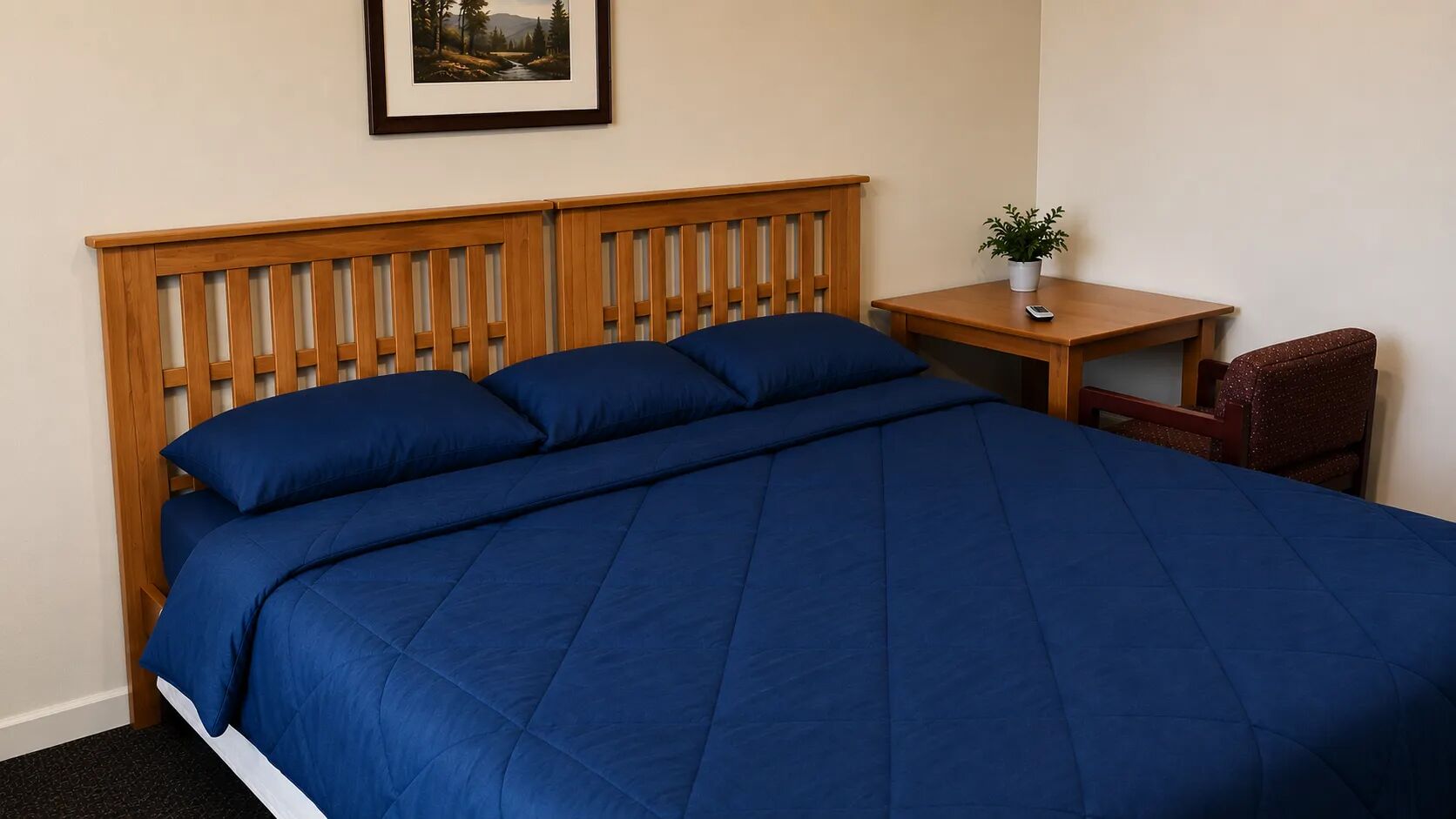 A hotel room with a wooden headboard, a large bed with blue bedding, a small table with a plant, and a chair.