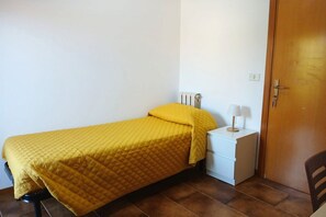 Ein kleines Zimmer mit einem Einzelbett, einer gelben Tagesdecke, einem Nachttisch mit Lampe und einer Tür rechts.