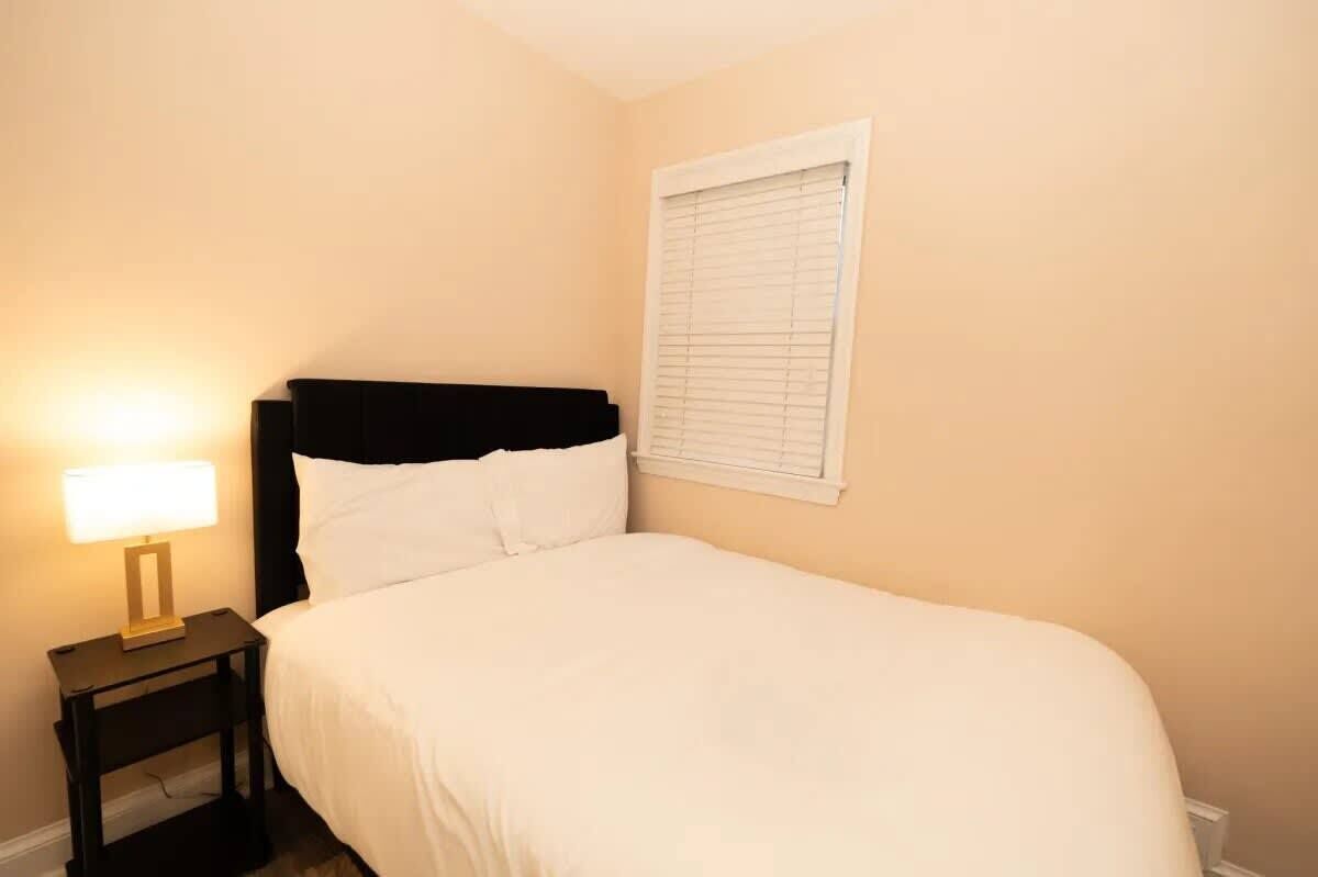 A single bed with a black headboard in a room with beige walls and a window with blinds.