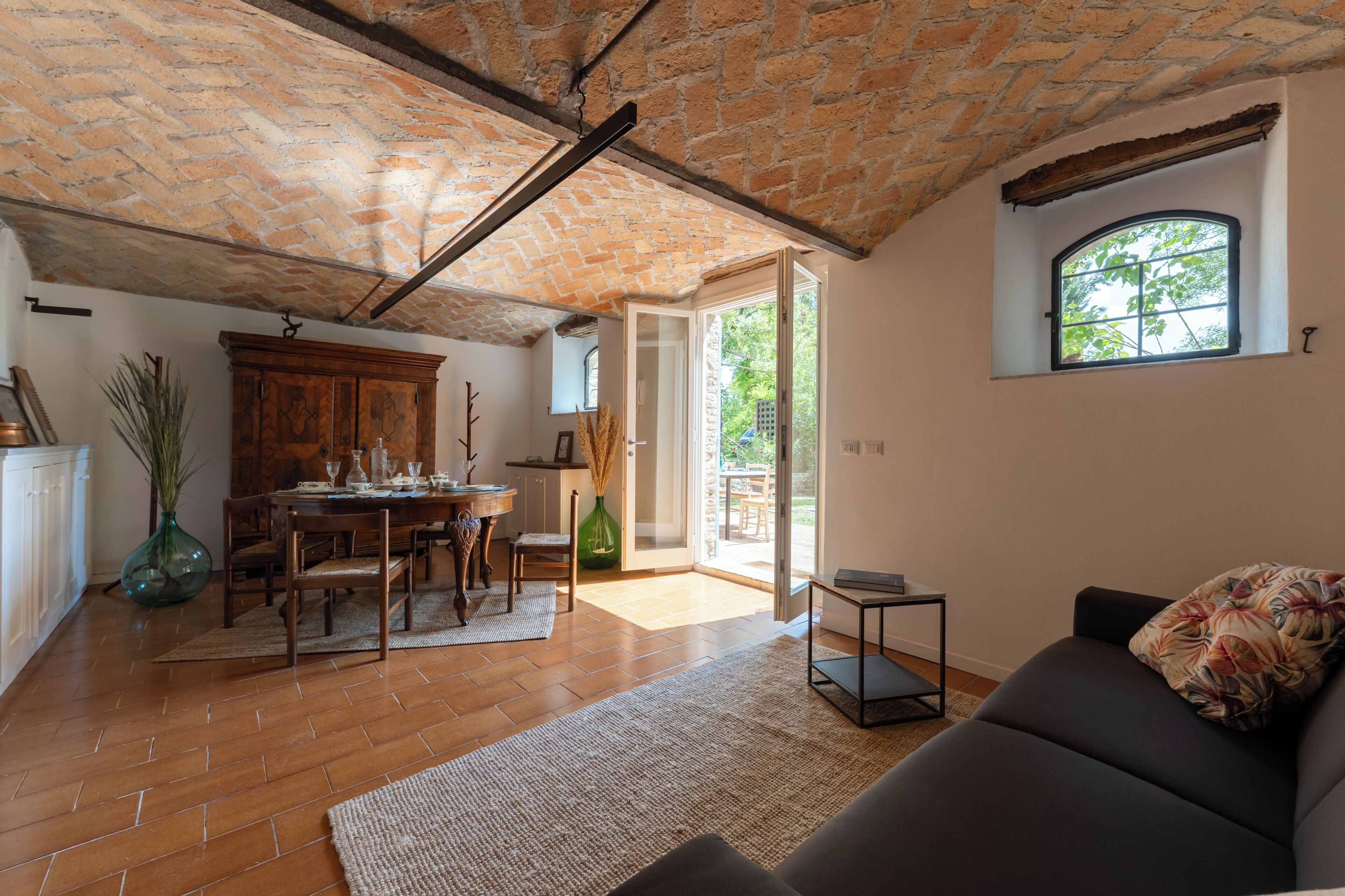 A spacious living area with a brick ceiling, a dining table set with chairs, a sofa, and a window with a view of greenery.