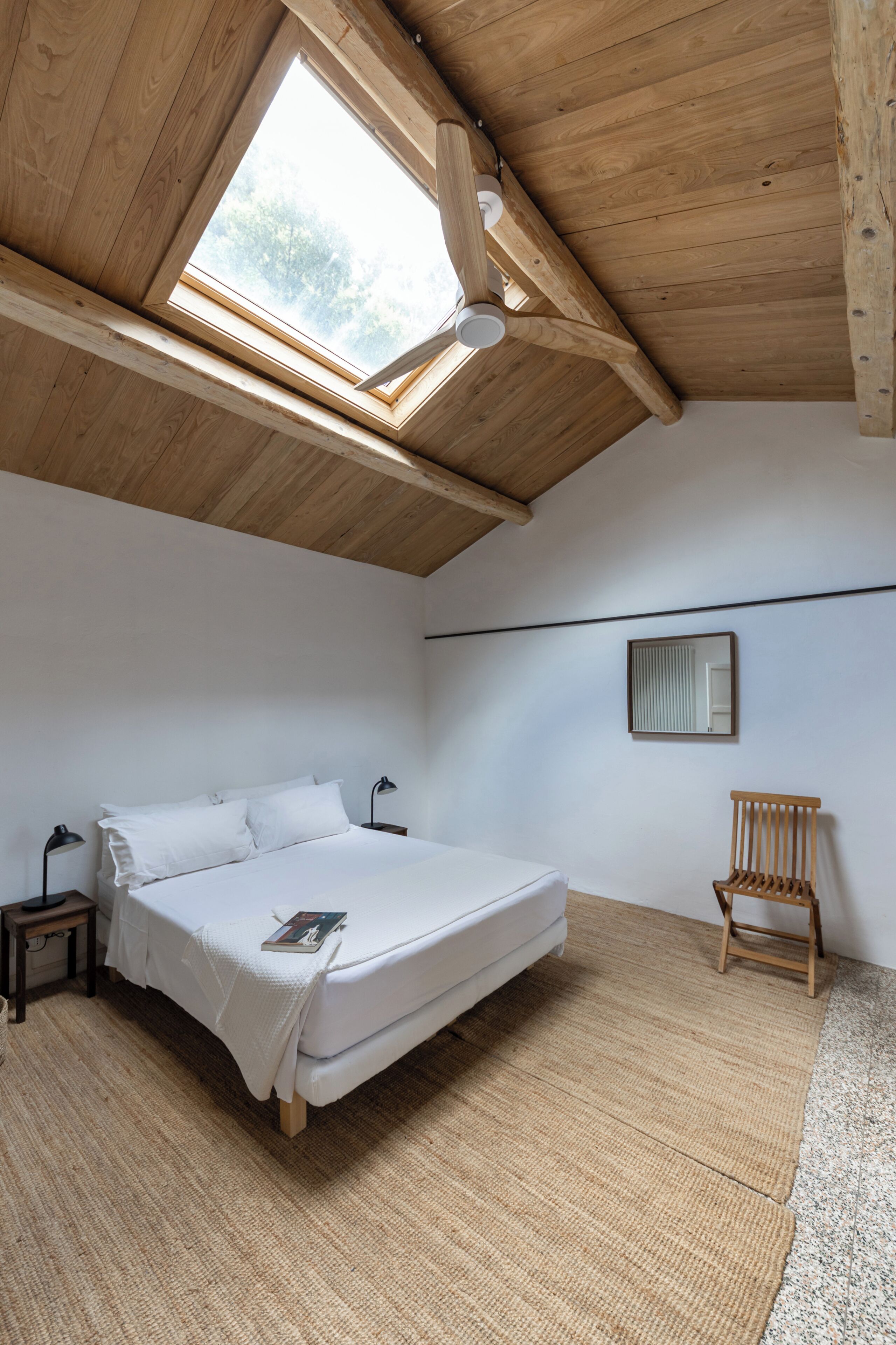 Ein Schlafzimmer mit schrägem Dach, einem Bett, einem Sessel, einem Nachttisch und einem Fenster.