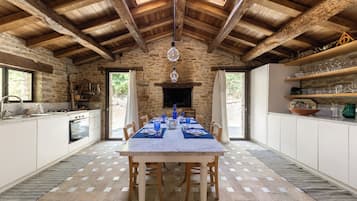 A spacious kitchen with a large dining table set for six, stone walls, wooden beams, and a view through the open door.
