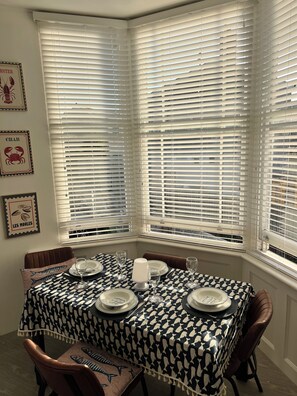 Un coin repas avec une table dressée pour quatre personnes, des stores blancs aux fenêtres et des tableaux décoratifs sur le thème des fruits de mer.