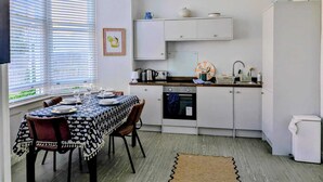 Cocina compacta con gabinetes blancos, una mesa de comedor para cuatro personas y un ventanal con persianas.