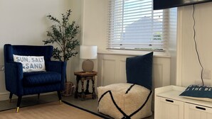 Una habitación con un sillón azul, una mesita con lámpara, un armario blanco y un ventanal grande con persianas.