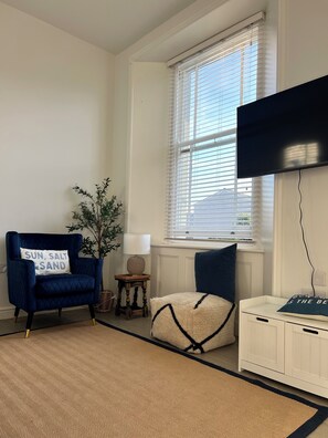 A room with a blue armchair, a small table with a lamp, a white cabinet, and a large window with blinds. - Jurassic coast spacious and stylish 2 bed 2 bath apartment. With own parking. (Portland)