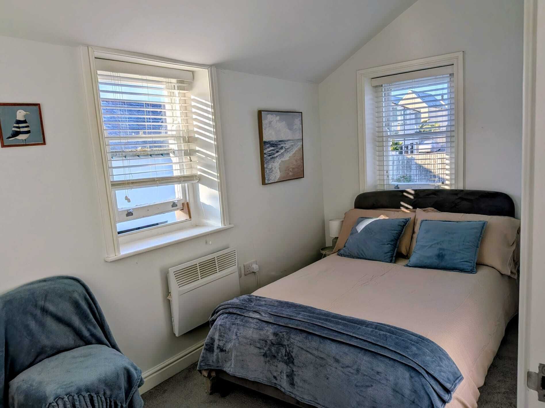 A bedroom with a bed, two windows with blinds, a radiator, a framed picture, and a chair.