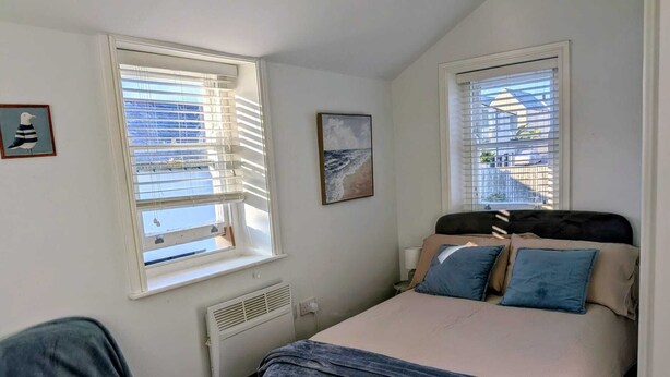Un dormitorio con una cama, dos ventanas con persianas, un radiador, un cuadro enmarcado y una silla.