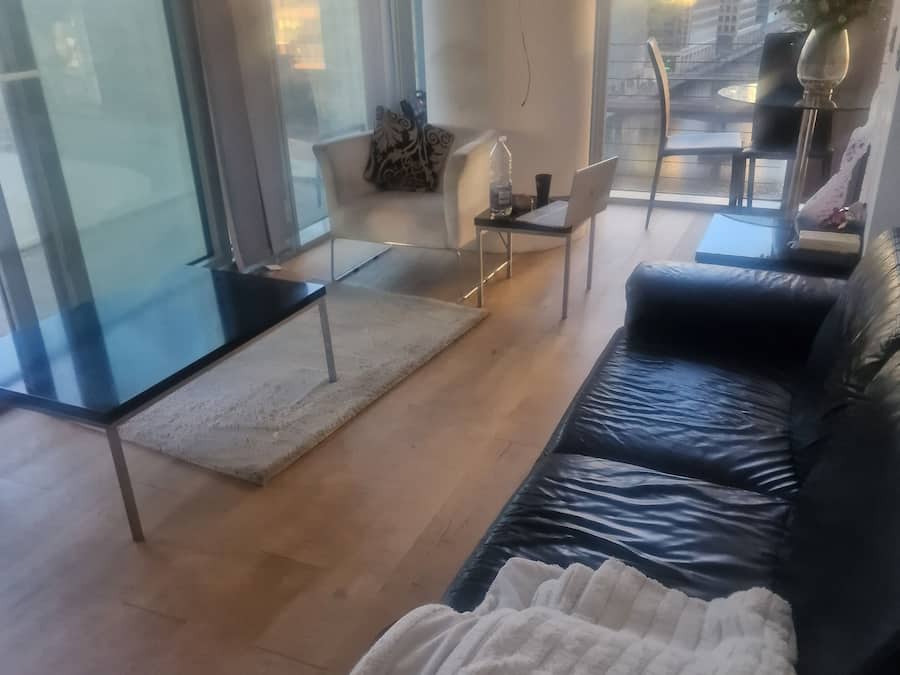 Una sala de estar moderna con un sofá de cuero negro, un separador de vidrio, un sillón blanco y una pequeña mesa con una computadora portátil.