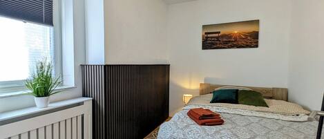 Un dormitorio con cama, radiador, ventana con persianas, una planta, una lámpara de pared y un cuadro.