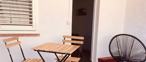 A patio with a wooden table and chairs, a black woven chair, and a window with blinds.