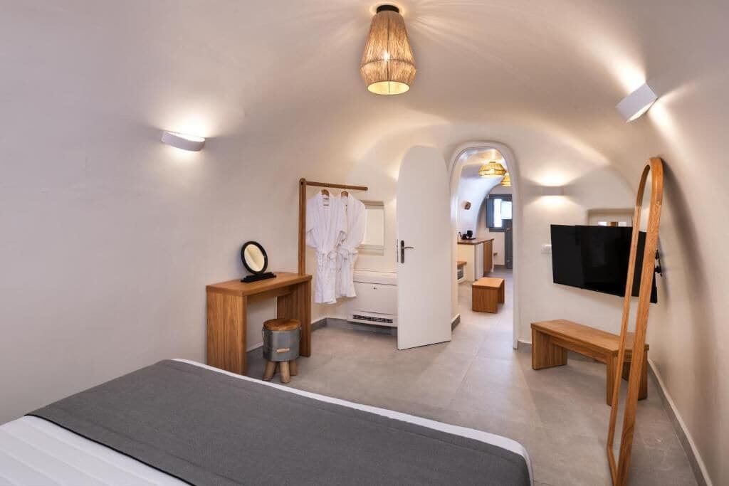A modern hotel room with a bed, a wooden side table, a mirror, a robe, a television, and a bathroom visible through an open door.