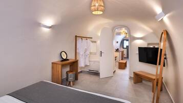 A modern hotel room with a bed, a wooden side table, a mirror, a robe, a television, and a bathroom visible through an open door.