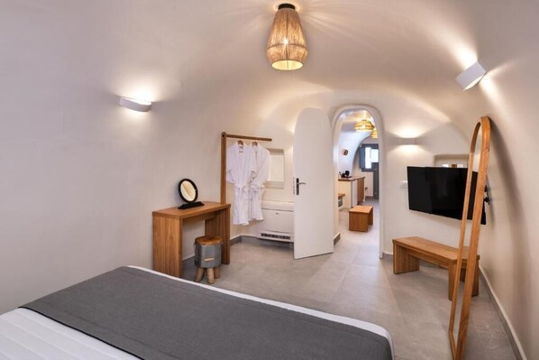 Habitación de hotel moderna con una cama, una mesita de madera, un espejo, una bata, un televisor y un baño visible a través de una puerta abierta.