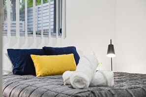 A neatly made bed with a grey quilt, two pillows in blue and yellow, and a rolled white towel. - Apartment - Ground Floor Sydney (Parramatta)