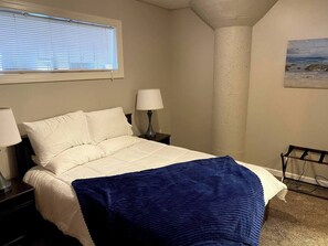 Ein Schlafzimmer mit einem Bett, zwei Nachttischlampen, einem Fenster mit Jalousien, einem Bild an der Wand und einem Sessel.