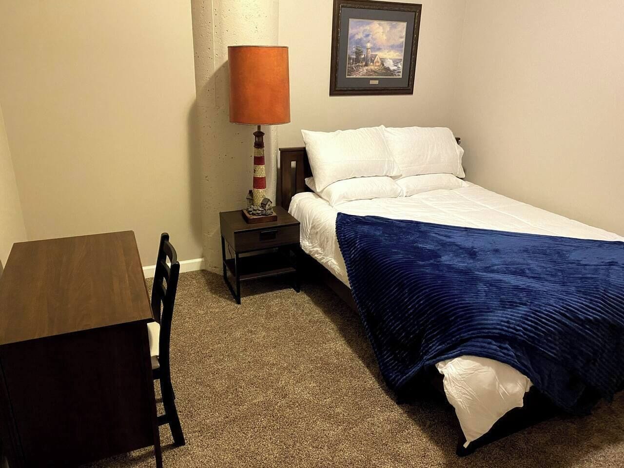 Un dormitorio pequeño y ordenado con una cama individual, una mesita de noche, una mesa y una lámpara.