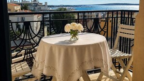 Ein Balkon mit rundem Tisch und Stühlen, einer Blumenvase und Blick auf das Meer sowie Gebäude.
