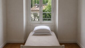 Ein kleines Zimmer mit einem Einzelbett, Holzboden, einem weißen Bett und einem Fenster mit Blick auf einen Garten.