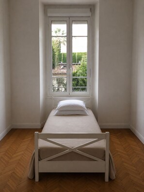 A small, single-bed room with a wooden floor, a white bed, and a window overlooking a garden. - Charming 2-bedroom apartment with AC in beautiful Cannes (Cannes)